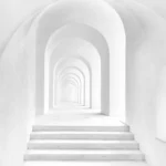 Minimalist white hallway with symmetrical archways and steps leading forward, creating a tunnel-like effect with strong perspective and soft natural lighting.